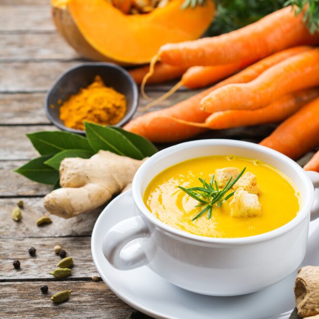 Carrot Squash and Ginger Soup served in a white bowl with handle. Carrots and Ginger are presented next to the bowl.